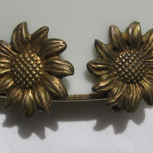 以下が含まれることがあります： ゴールドトーンのひまわりイヤリングのペア。イヤリングは金属製で、詳細なひまわりのデザインが施されています。