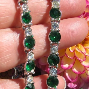 May include: A silver-toned tennis bracelet featuring a repeating pattern of round, faceted emerald green stones alternating with smaller, clear, round, faceted stones set in a metallic frame, creating a continuous line of sparkling jewelry.