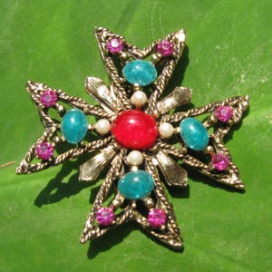 Puede incluir: Broche vintage dorado en forma de cruz, adornado con una piedra roja central, cabujones turquesa y peque&ntilde;os brillantes rosas. El broche presenta un dise&ntilde;o texturizado detallado con perlas de imitaci&oacute;n, sobre un fondo verde.