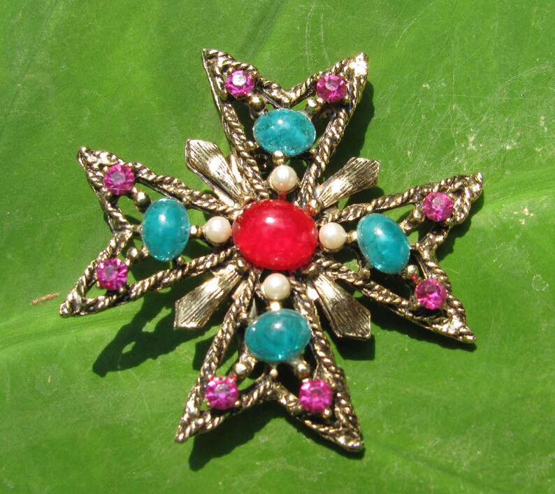 Puede incluir: Broche vintage dorado en forma de cruz, adornado con una piedra roja central, cabujones turquesa y peque&ntilde;os brillantes rosas. El broche presenta un dise&ntilde;o texturizado detallado con perlas de imitaci&oacute;n, sobre un fondo verde.