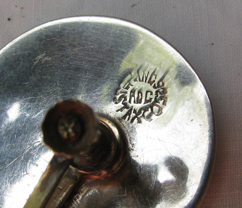 Puede incluir: Primer plano de un objeto de metal plateado con un sello circular que dice "STERLING 925 MEXICO".