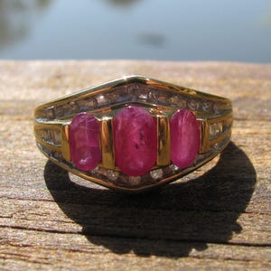 Op de afbeelding: Een gouden ring met drie ovale, levendige rode edelstenen. De ring heeft een gouden band met kleinere, heldere stenen aan de zijkanten. De ring wordt gepresenteerd op een houten oppervlak, met een vage achtergrond.