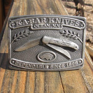 May include: A rectangular silver-colored belt buckle featuring a detailed knife design. The buckle has the text "KABAR KNIVES OLEAN, N.Y." at the top and "DEPENDABLE SINCE 1898" at the bottom. A knife is in the center.