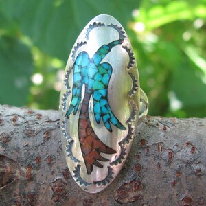 May include: A silver ring with an oval-shaped turquoise and red stone inlay. The inlay is a stylized design of a bird with outstretched wings.
