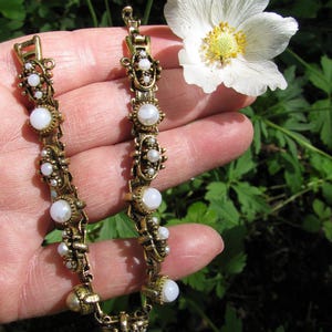 May include: A vintage gold-toned bracelet with white, pearl-like accents. The bracelet is displayed against a backdrop of green foliage and a white flower with a yellow center. The bracelet's design features ornate detailing and a clasp.