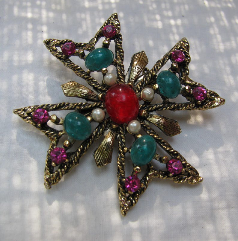 Puede incluir: Broche vintage en forma de estrella, con un marco de metal dorado. El broche presenta una piedra roja central, rodeada de piedras preciosas verdes y rosas, y peque&ntilde;as perlas blancas. El dise&ntilde;o de estrella est&aacute; detallado con bordes en forma de cuerda.