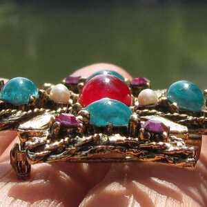 Puede incluir: Broche vintage dorado con dise&ntilde;o de cruz. Presenta una piedra roja central, rodeada de piedras turquesas y moradas, y peque&ntilde;as perlas blancas. El broche tiene un trabajo de metal ornamentado y texturizado.