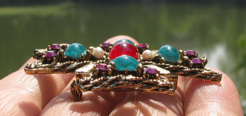Puede incluir: Broche vintage dorado con dise&ntilde;o de cruz. Presenta una piedra roja central, rodeada de piedras turquesas y moradas, y peque&ntilde;as perlas blancas. El broche tiene un trabajo de metal ornamentado y texturizado.