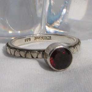 May include: A silver ring featuring a round, deep red garnet gemstone set in a bezel. The band has a repeating triangular pattern along its outer surface. The interior of the band is stamped with the text 925 and a hallmark.
