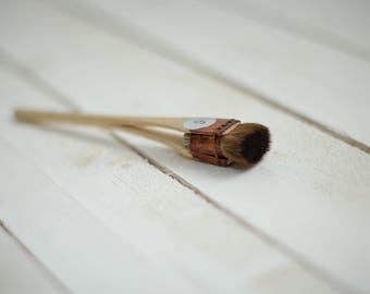 May include: A close-up of a small, brown paintbrush with a wooden handle and a white label with the number "8" on it. The brush is lying on a white wooden surface.