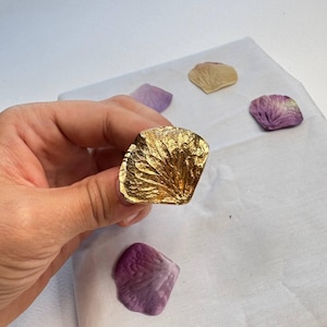 May include: A gold-toned ring featuring a textured, organic petal-shaped design that mimics the veins and contours of a natural flower petal. The surface has a metallic, reflective finish with deep ridges and fine details across the entire face.