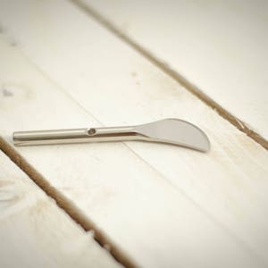 May include: A silver metal tool with a curved blade and a handle. The tool is lying on a white wooden surface.