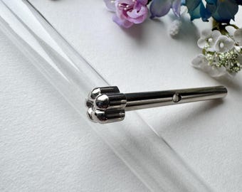 Può includere: Una provetta di vetro trasparente con un tappo color argento. Il tappo ha un design decorativo a forma di fiore. La provetta è posizionata in diagonale, con il tappo in alto. Fiori artificiali nei toni del viola e del blu sullo sfondo.