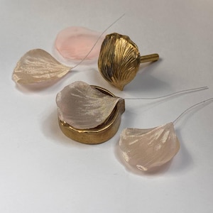 Può includere: Petali di fiori artificiali e un bocciolo di fiore dorato. I petali sono in tessuto trasparente nei toni del rosa e beige, attaccati a sottili fili di ferro. Il bocciolo è una scultura dorata dettagliata.