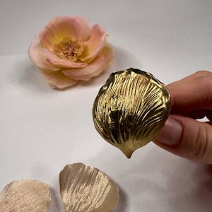 May include: A gold-coloured, textured, petal-shaped ring is held in a hand. A peach-coloured flower and two petals are in the background. The ring has a pointed tip and a ridged surface.