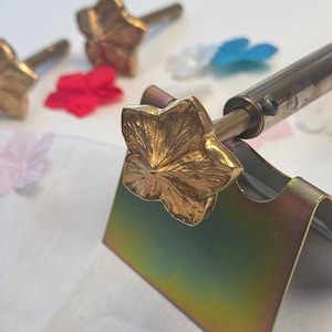Könnte beinhalten: Ein goldfarbener, blumenförmiger Brennstempelkopf aus Metall an einem Metallstab, der auf einem Metallständer ruht. Zwei weitere identische blumenförmige Metallköpfe sind im Hintergrund zu sehen, zusammen mit verstreuten kleinen Stoffblütenblättern.