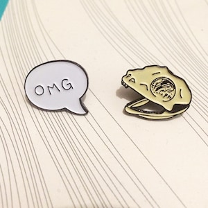 Pode incluir: Dois pinos de esmalte, um branco com o texto "OMG" em letras pretas e o outro um crânio amarelo com contorno preto.