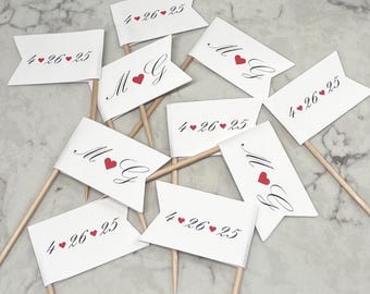 May include: A set of eight white flags with red hearts and the text "4.26.25" and "M & G" printed on them. The flags are attached to wooden skewers.
