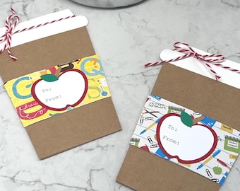 May include: Two brown paper coffee cup-shaped gift cards with white tops and red and white twine. Each card has a colourful band with an apple graphic and space for "To:" and "From:" messages. A succulent plant in a marble pot is in the background.