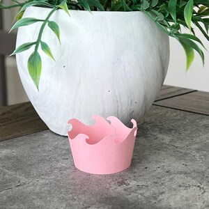 May include: A pink cupcake liner with a wave design. The liner is sitting on a gray surface in front of a white planter with green plants.