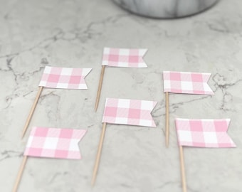 May include: Six pink and white gingham flag cupcake toppers on wooden sticks. A marble-effect pot with green plants is in the background. The flags are arranged on a white and grey marble surface.
