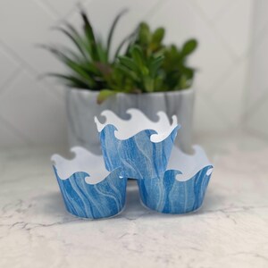 May include: Three blue and white cupcake liners with a wave design. The liners have a blue marbled pattern and a white wave-shaped top edge. A small potted succulent is in the background.