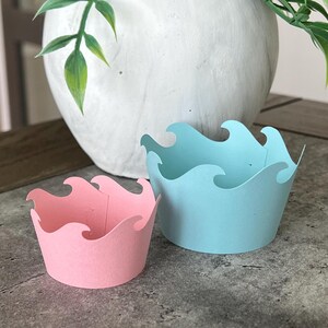 May include: Two cupcake liners in the shape of ocean waves. One is light blue and the other is pink.