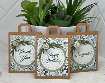 May include: Three brown paper gift bags with scalloped edges and handles. Each bag features a white circle with a message: "Thank You", "Happy Birthday", and "Congrats!". The bags are decorated with floral and leaf patterns and a small flower.