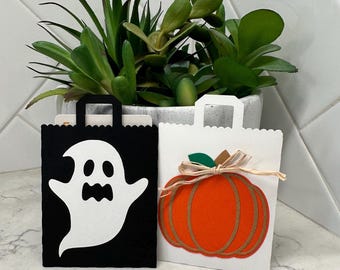 May include: Two Halloween-themed gift bags. One black bag features a white ghost design. The other is white with an orange pumpkin design, a green leaf, and a tan ribbon. Both bags have a scalloped top edge and a handle.