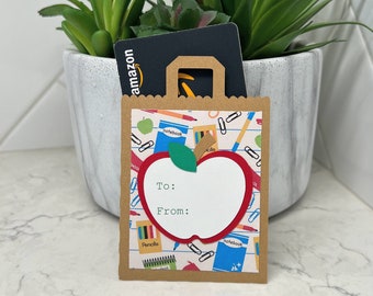 May include: A brown paper gift bag featuring a red apple design, a green leaf, and the words "To:" and "From:" on the front. The bag contains an Amazon gift card and is decorated with school-themed patterns. A potted succulent plant is in the background.