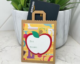 May include: A brown paper bag gift tag with a red apple design. The apple has a white centre with the words "To:" and "From:" written on it. The bag has a yellow and brown pattern with school-themed graphics. An Amazon gift card is tucked inside the bag.