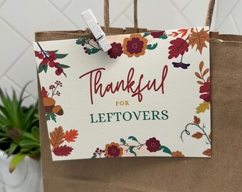 May include: A brown paper gift bag with a tag that reads "Thankful for Leftovers." The tag features autumnal floral and leaf illustrations in red, orange, and green. A small white clothes peg secures the tag to the bag. The bag has twisted paper handles.