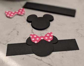 May include: Black and pink paper cutouts of Mickey Mouse and Minnie Mouse heads with polka dot bows. The cutouts are on a white marble surface.