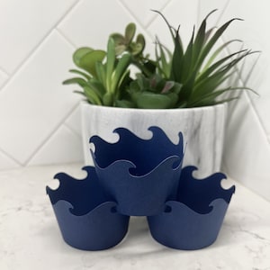 May include: Three navy blue cupcake liners with a wave-like design. The liners are stacked in front of a white marble planter with green succulents. The liners are made of paper and are designed to hold cupcakes or muffins.