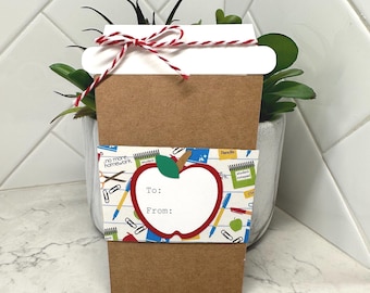 May include: A brown paper coffee cup with a white lid tied with red and white twine. A decorative band with an apple design and the words "To:" and "From:" wraps around the cup. The cup is a gift for a teacher.