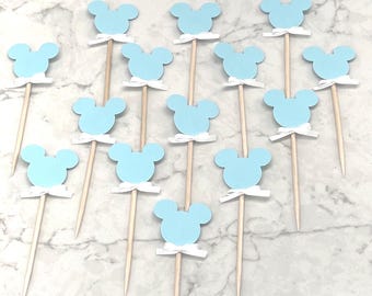 May include: A set of 15 blue and white Mickey Mouse themed cupcake toppers. Each topper features a light blue Mickey Mouse head with white ears and a white bow.