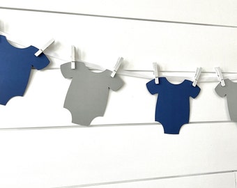 May include: A string of blue and grey paper baby onesies hanging on a white wooden background with white clothes pegs.