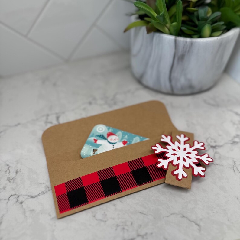 May include: A brown gift card holder with a red and black buffalo plaid design. A gift card with a snowman design is partially visible. A white and red snowflake ornament is attached to the holder, suggesting a winter or holiday theme.
