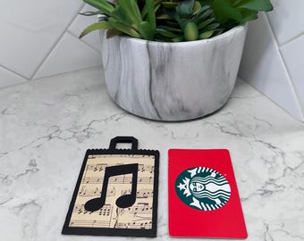 May include: A black gift tag featuring a musical note cutout, set against vintage sheet music. A red Starbucks gift card with a green logo is next to the tag. A potted succulent plant is in the background.