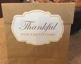 May include: A brown paper bag with a white and orange label that reads "Thankful FOR LEFTOVERS". The bag is folded at the top. A black basket filled with corks is in the background. The text is in an elegant script.
