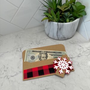 May include: A brown paper gift card holder with a red and black buffalo plaid ribbon and a white snowflake. A twenty-dollar bill is partially visible inside the holder. A small potted succulent is in the background.