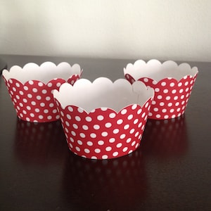 May include: Three red cupcake liners with white polka dots. The liners have a scalloped edge and are arranged on a dark surface. The interior of the liners is white, providing a contrast to the red and white exterior.