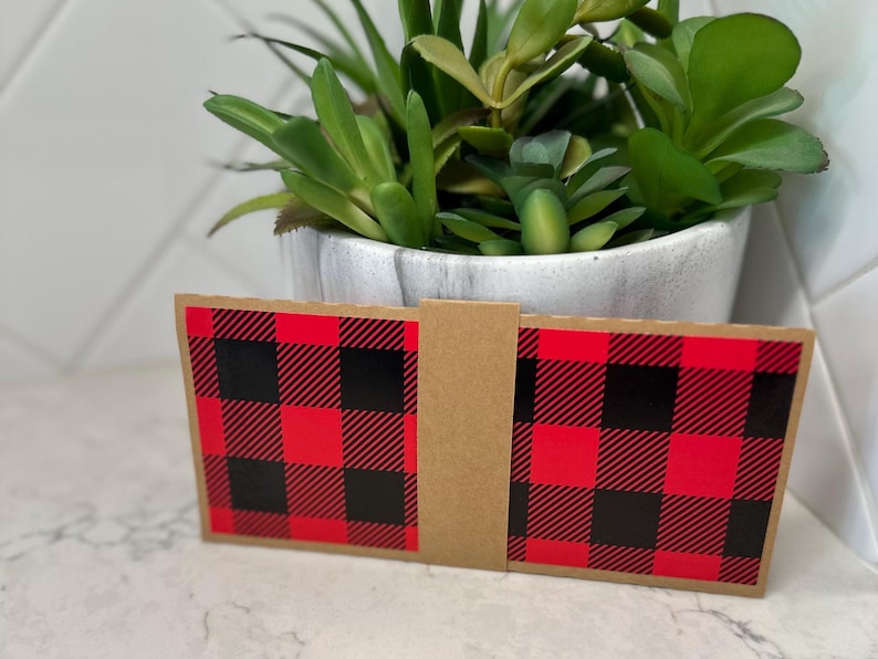 May include: A red and black buffalo plaid design on a folded cardboard item. The item is in front of a white pot with a green plant. The plaid pattern is a classic design.