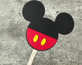 May include: A black and red Mickey Mouse head-shaped decoration with two yellow ovals, attached to a wooden stick. This item is likely for a party or cake topper, featuring a classic cartoon character.