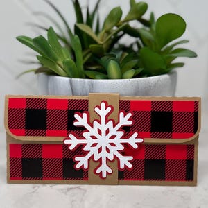 May include: A small, rectangular gift box with a red and black buffalo plaid pattern. A white snowflake is attached to the front. The box is made of cardboard and has a flap closure. A small potted plant is in the background.