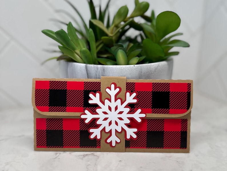 May include: A small, rectangular gift box with a red and black buffalo plaid pattern. A white snowflake is attached to the front. The box is made of cardboard and has a flap closure. A small potted plant is in the background.