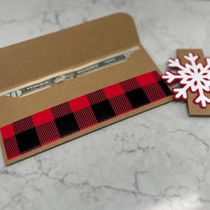 May include: A brown paper money holder with a red and black buffalo plaid design. A white and red snowflake decoration is next to the holder. A twenty-dollar bill is visible inside the holder.