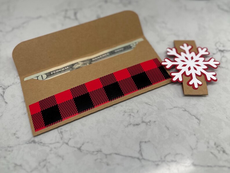 May include: A brown paper money holder with a red and black buffalo plaid design. A white and red snowflake decoration is next to the holder. A twenty-dollar bill is visible inside the holder.