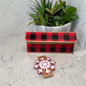 May include: A rectangular gift box with a red and black plaid pattern. A white and red snowflake decoration is in front of the box. A small potted succulent plant is in the background.