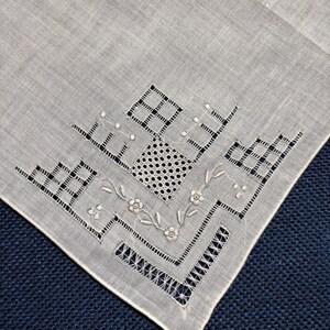 May include: A white linen handkerchief featuring intricate white-on-white cutwork embroidery. The corner design includes geometric grid patterns, small circular eyelet details, and a delicate floral vine motif with stitched leaves and petals.